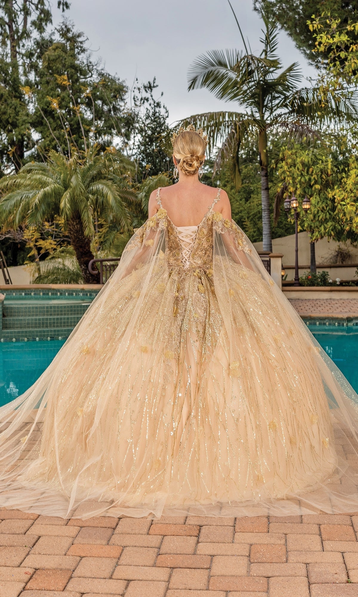 A woman stands by a pool, surrounded by palm trees and lush greenery, wearing the Quinceanera Dress 1830 By Dancing Queen—a gold gown with off-the-shoulder cape sleeves.